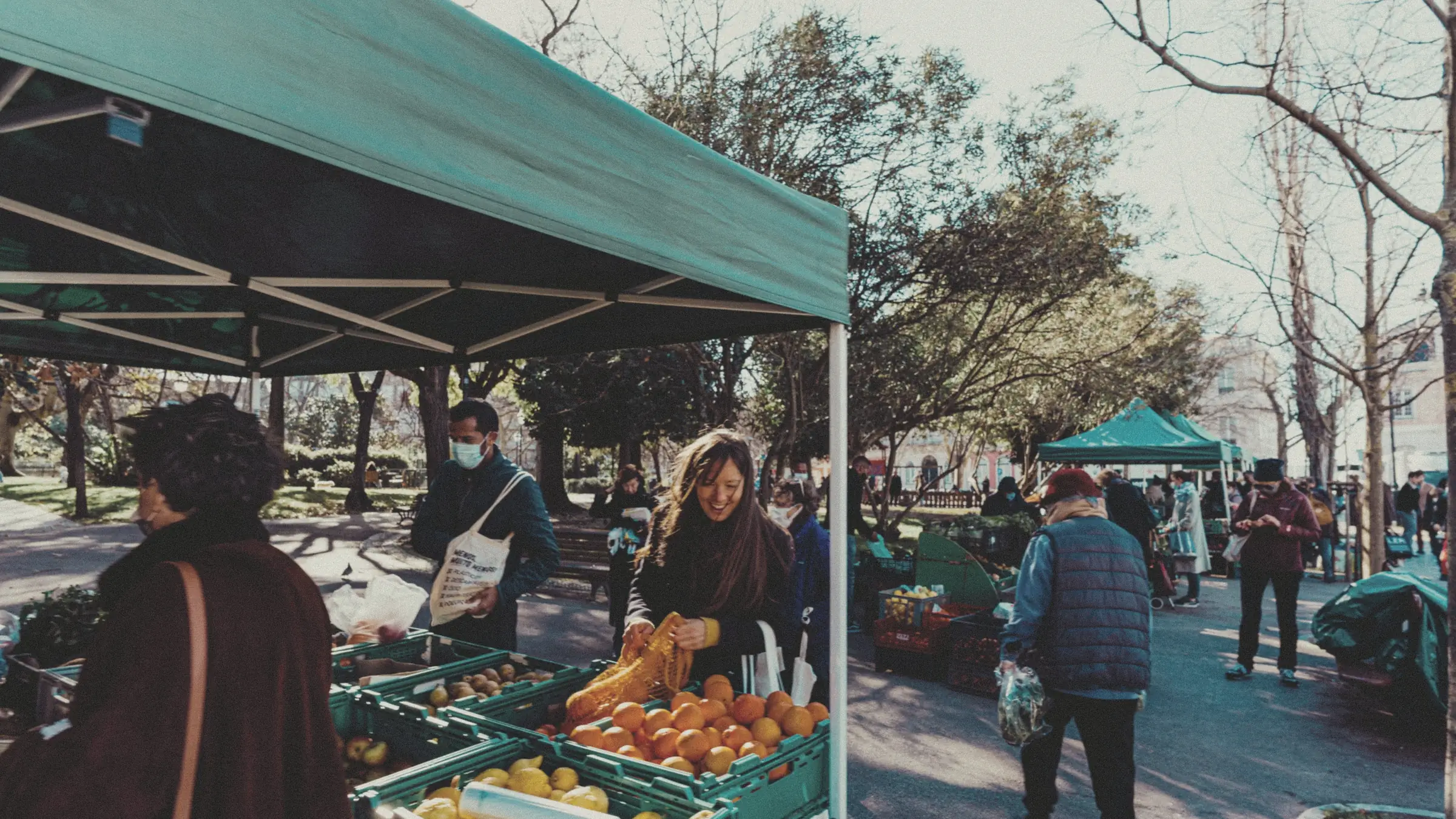 Shoppers and stallholders at an open-air farmers market under green awnings in Lisbon.