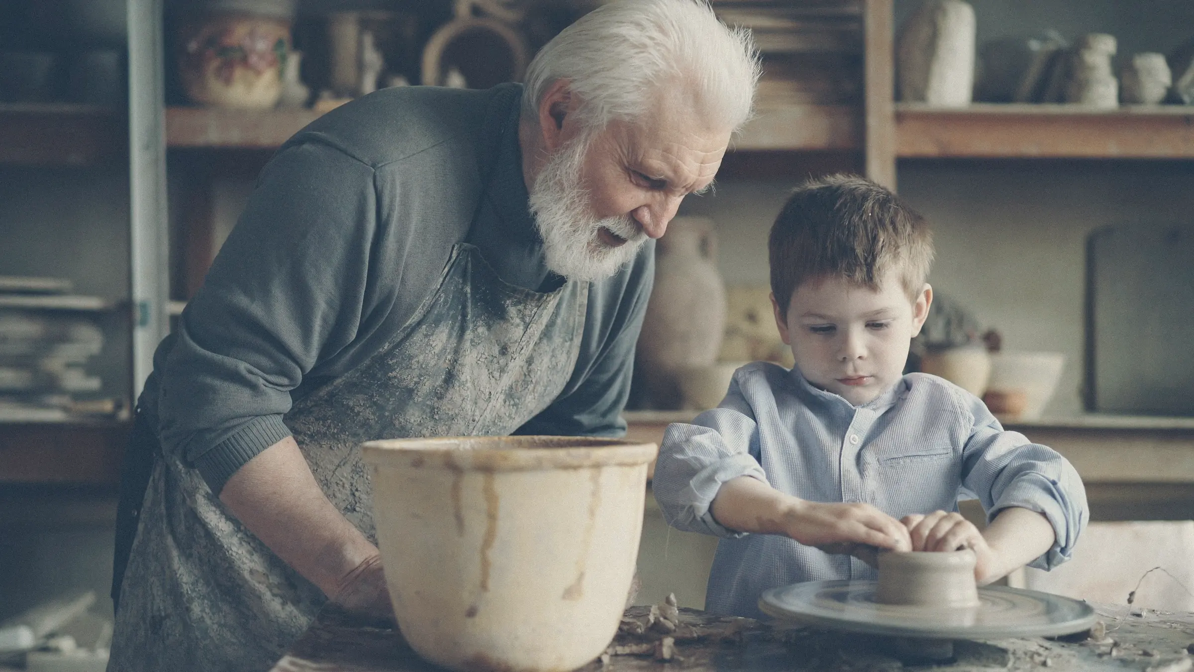 A white-bearded potter shows a young boy how to shape clay in a workshop lined with pots.