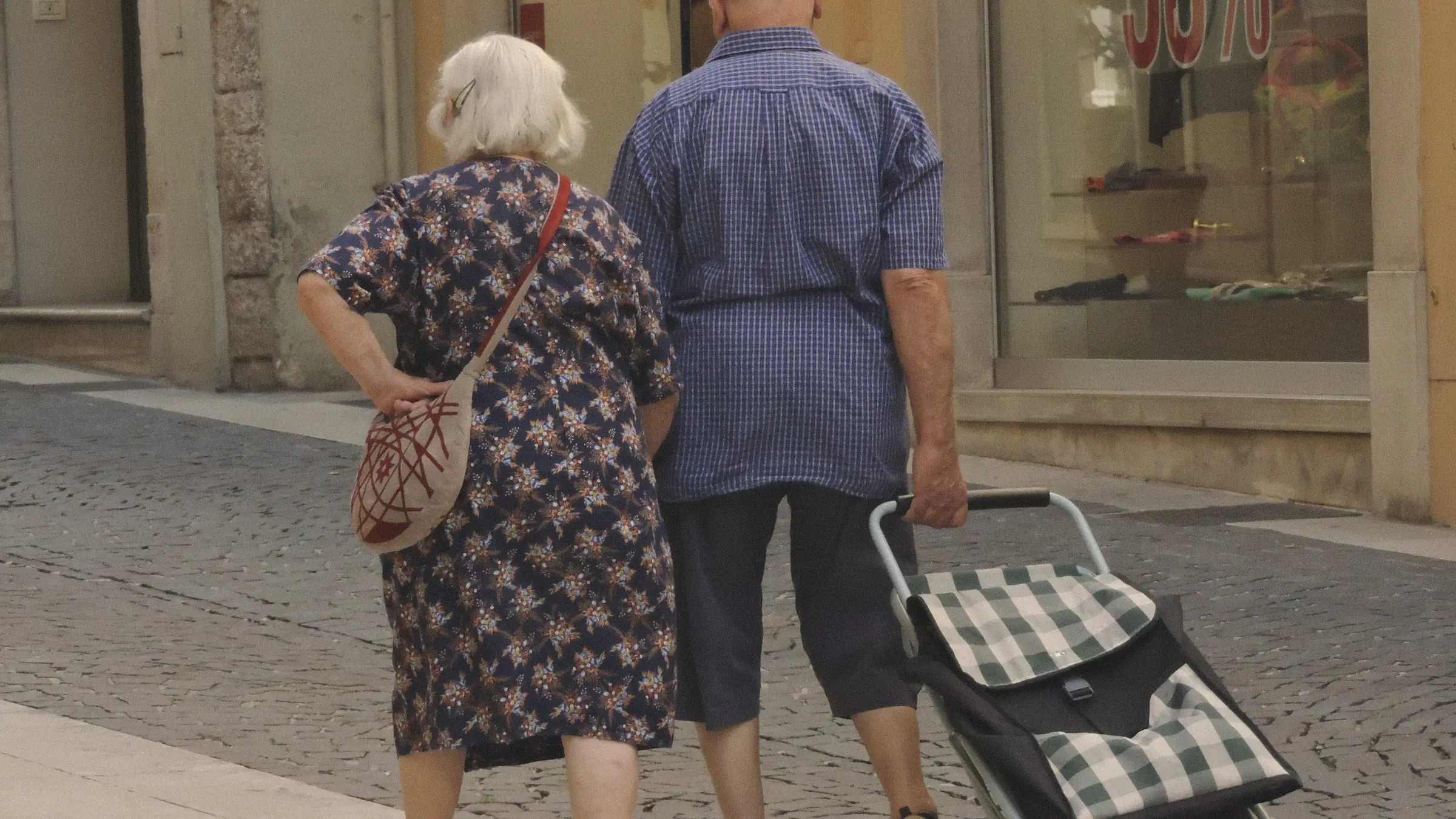 An older couple seen from behind walking down a city street, one pulling a shopping trolley.