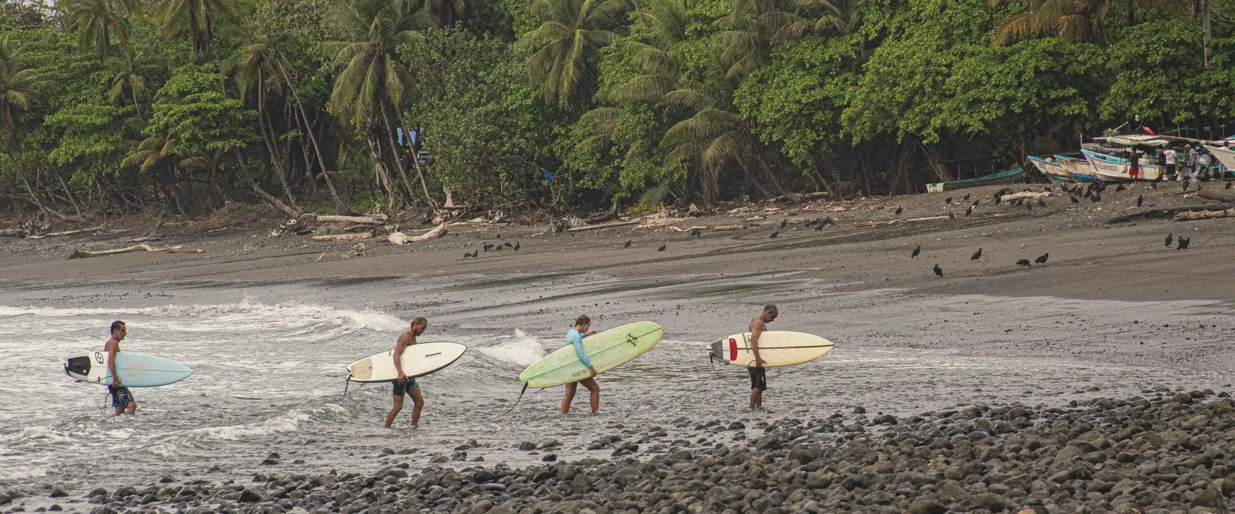 Surfers carrying their boards across a dark-sand beach into the Pacific at Pavones.