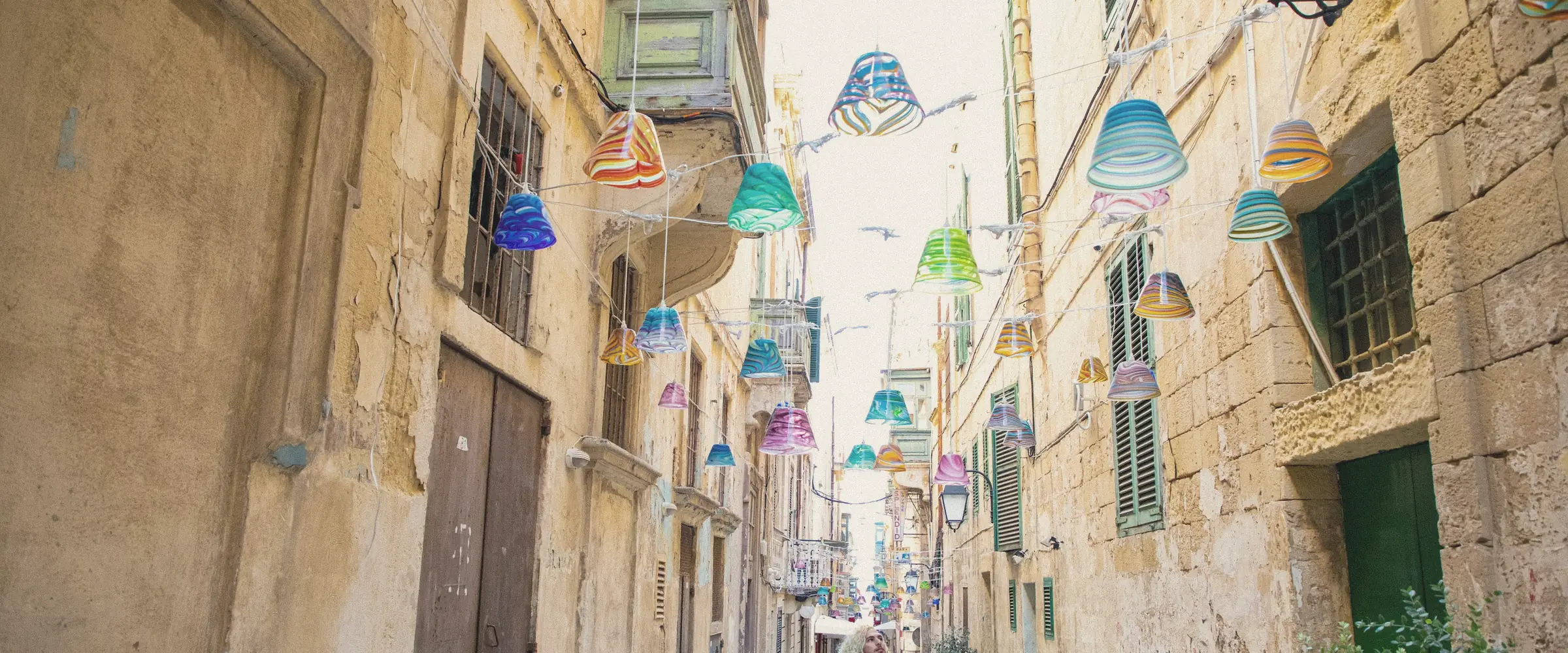 A narrow honey-coloured limestone street with green shutters and colourful lanterns strung overhead.