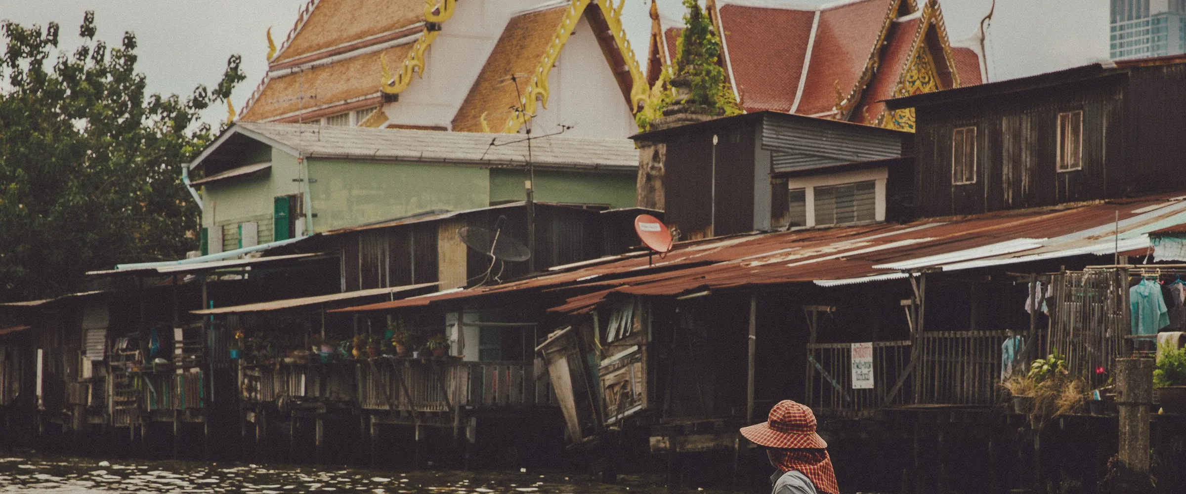 A woman paddles a small boat along a calm Bangkok canal in front of wooden houses.