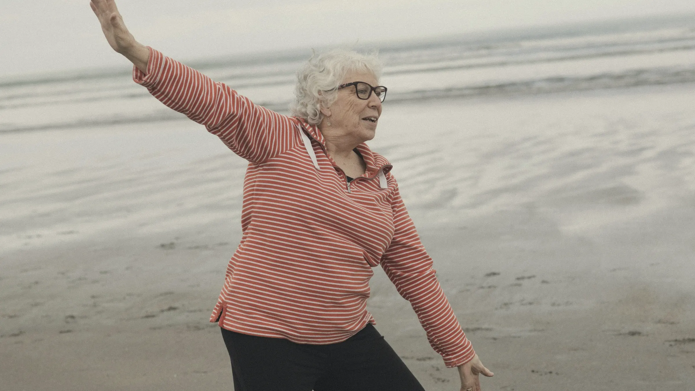 An older woman in a striped top stretches her arms out while moving on a misty beach.